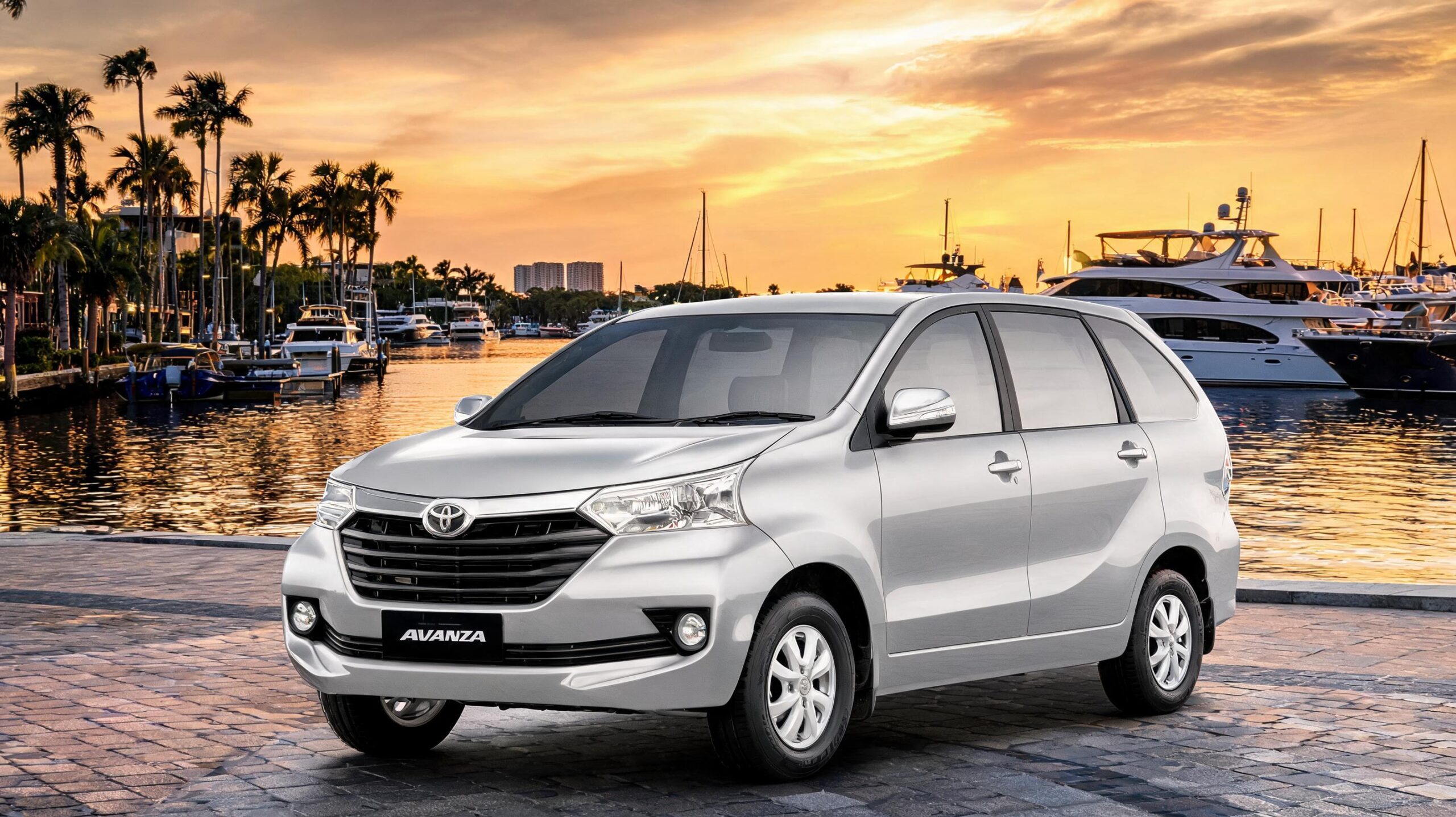 Mobil Toyota Avanza berwarna perak diparkir di tepi laut saat matahari terbenam, dengan kapal pesiar dan pohon palem di latar